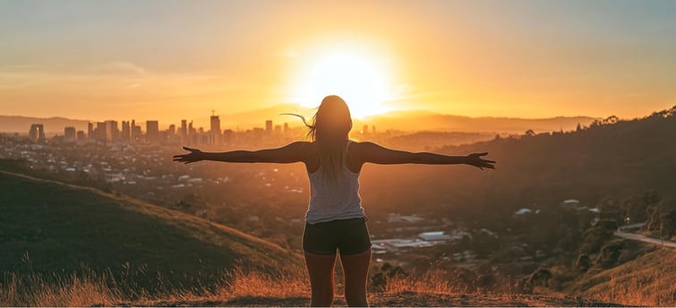 A woman with her arms outstretched on top of a hill overlooking city skyline at sunset.